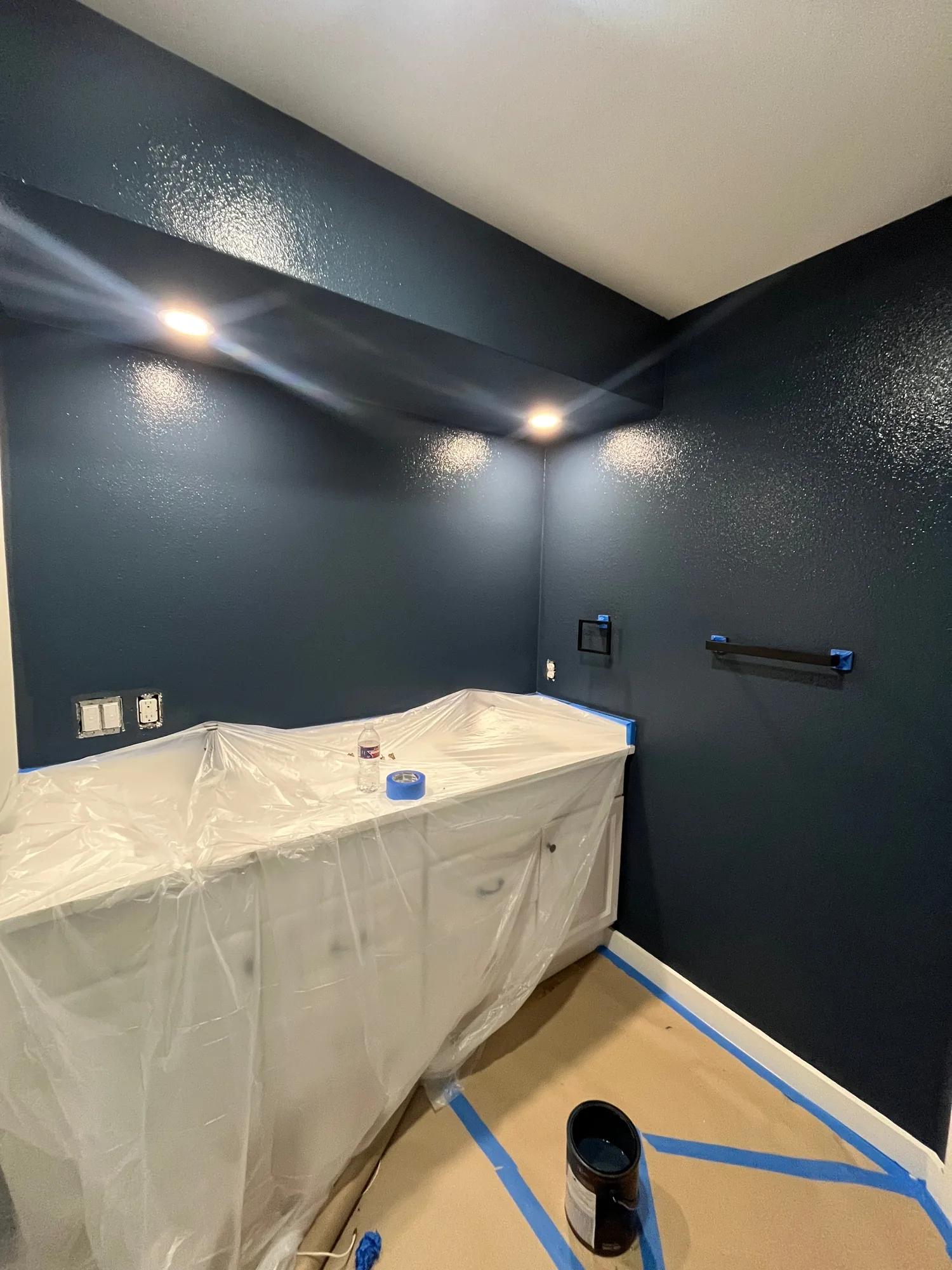 Bathroom vanity covered in plastic beneath dark blue walls with masking tape and protected flooring.